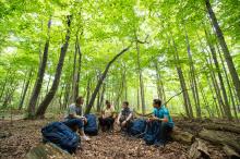 Photograph of four people sitting in a forest with hiking gear.